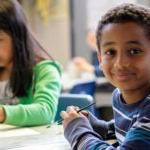 Two 学生 sit at a table in a classroom; one student smiles for a photo